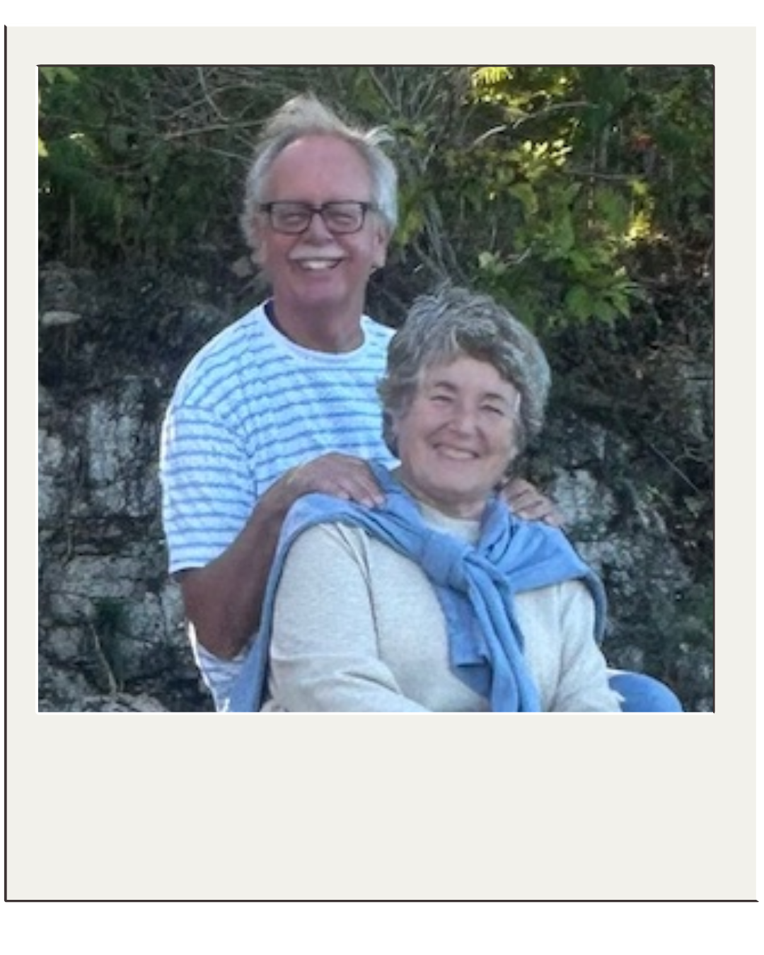 Polaroid Photo of man and woman in an outdoor setting with greenery and rocks.  The man wearing a striped shirt is standing behind the woman who is wearing a blue sweater tied around her shoulders and a cream colored shirt.  Both are smiling.